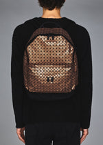 A person in black attire is seen from behind carrying the BAO BAO ISSEY MIYAKE DAYPACK REFLECTOR, a three-dimensional backpack featuring a brown and black geometric triangular grid design, against a plain gray background.