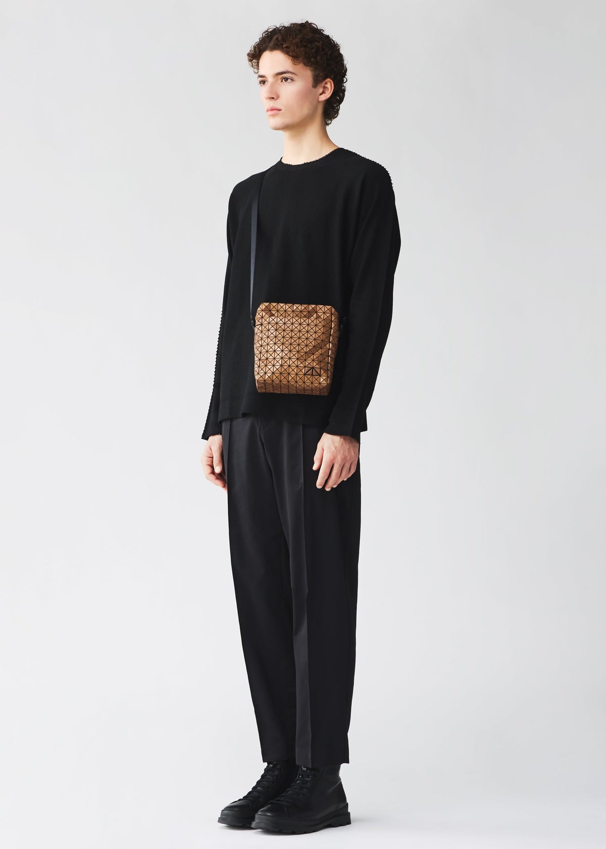 A person with curly hair wears black clothes and holds the BAO BAO ISSEY MIYAKE VOYAGER REFLECTOR tote, featuring a reflective surface and sleek black strap, standing against a plain light background for a stylish look. view 6 of 8