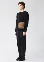 A person with curly hair wears black clothes and holds the BAO BAO ISSEY MIYAKE VOYAGER REFLECTOR tote, featuring a reflective surface and sleek black strap, standing against a plain light background for a stylish look.