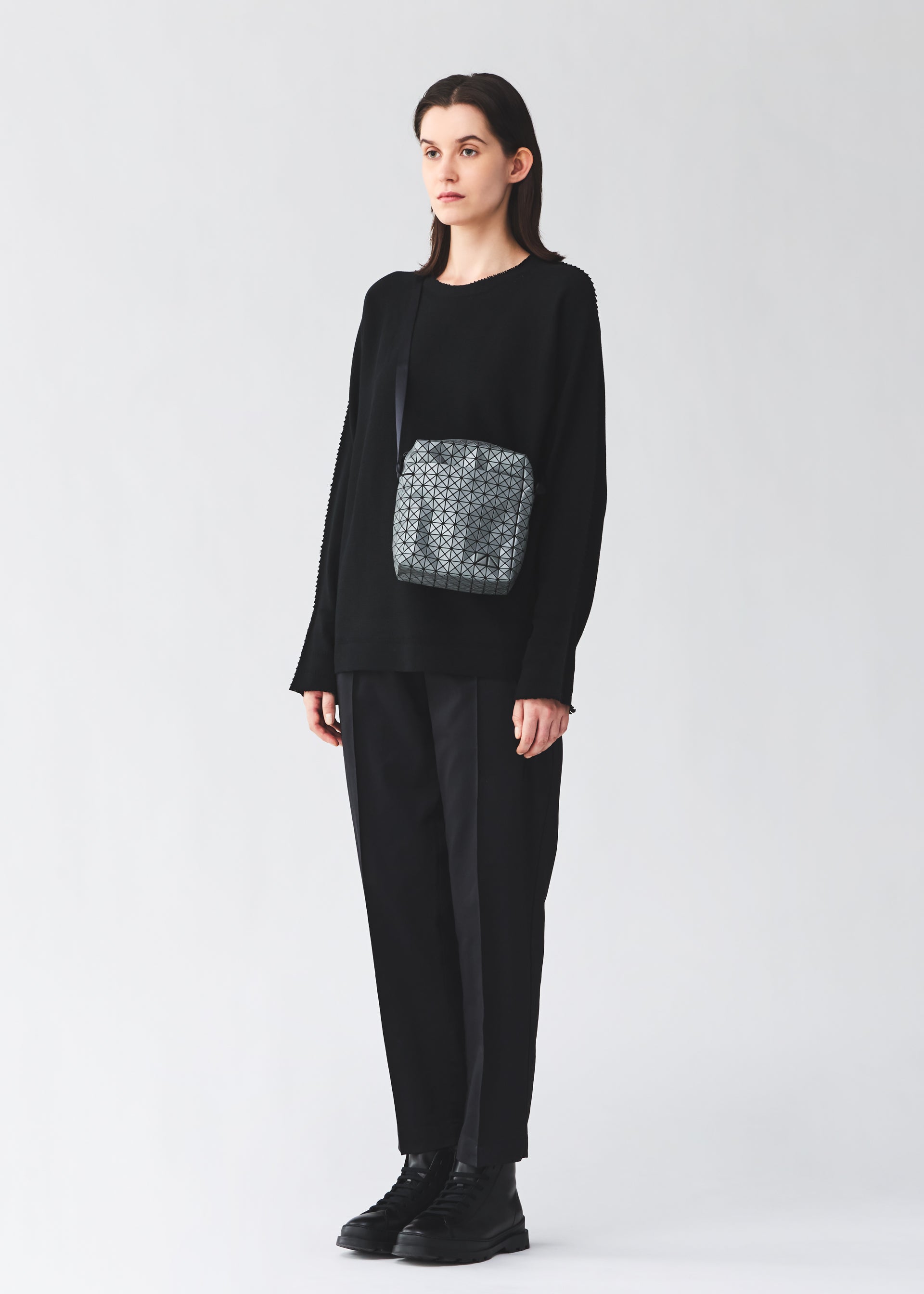 Zoomed view A person with long dark hair stands against a plain background, wearing a loose black sweater, striped black pants, black boots, a small geometric crossbody bag, and carrying the BAO BAO ISSEY MIYAKE VOYAGER REFLECTOR tote.