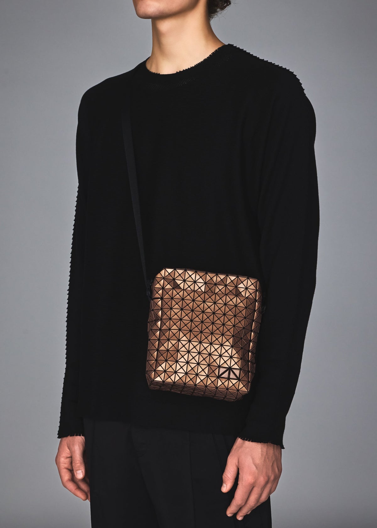 Wearing a black long-sleeve top and pants, a person carries the BAO BAO ISSEY MIYAKE VOYAGER REFLECTOR tote, a shiny metallic bag with geometric patterns and a black strap, against a plain gray background. Their head is partly out of frame. view 8 of 8