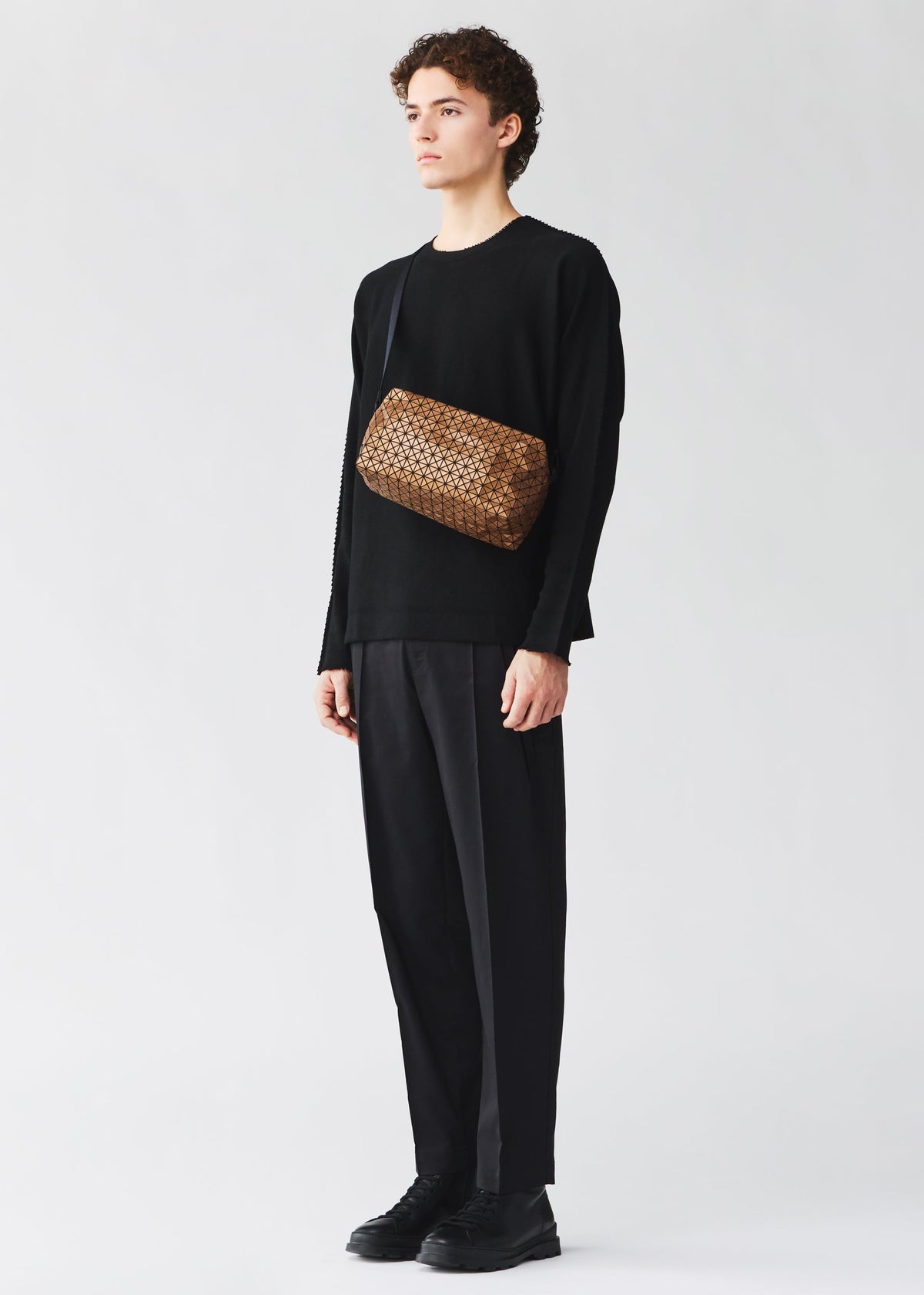 A young person with short curly hair, dressed in all black, stands facing left wearing the BAO BAO ISSEY MIYAKE SADDLE BAG REFLECTOR over one shoulder. The background is plain light gray. view 6 of 8