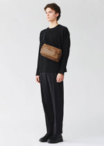 A young person with short curly hair, dressed in all black, stands facing left wearing the BAO BAO ISSEY MIYAKE SADDLE BAG REFLECTOR over one shoulder. The background is plain light gray.