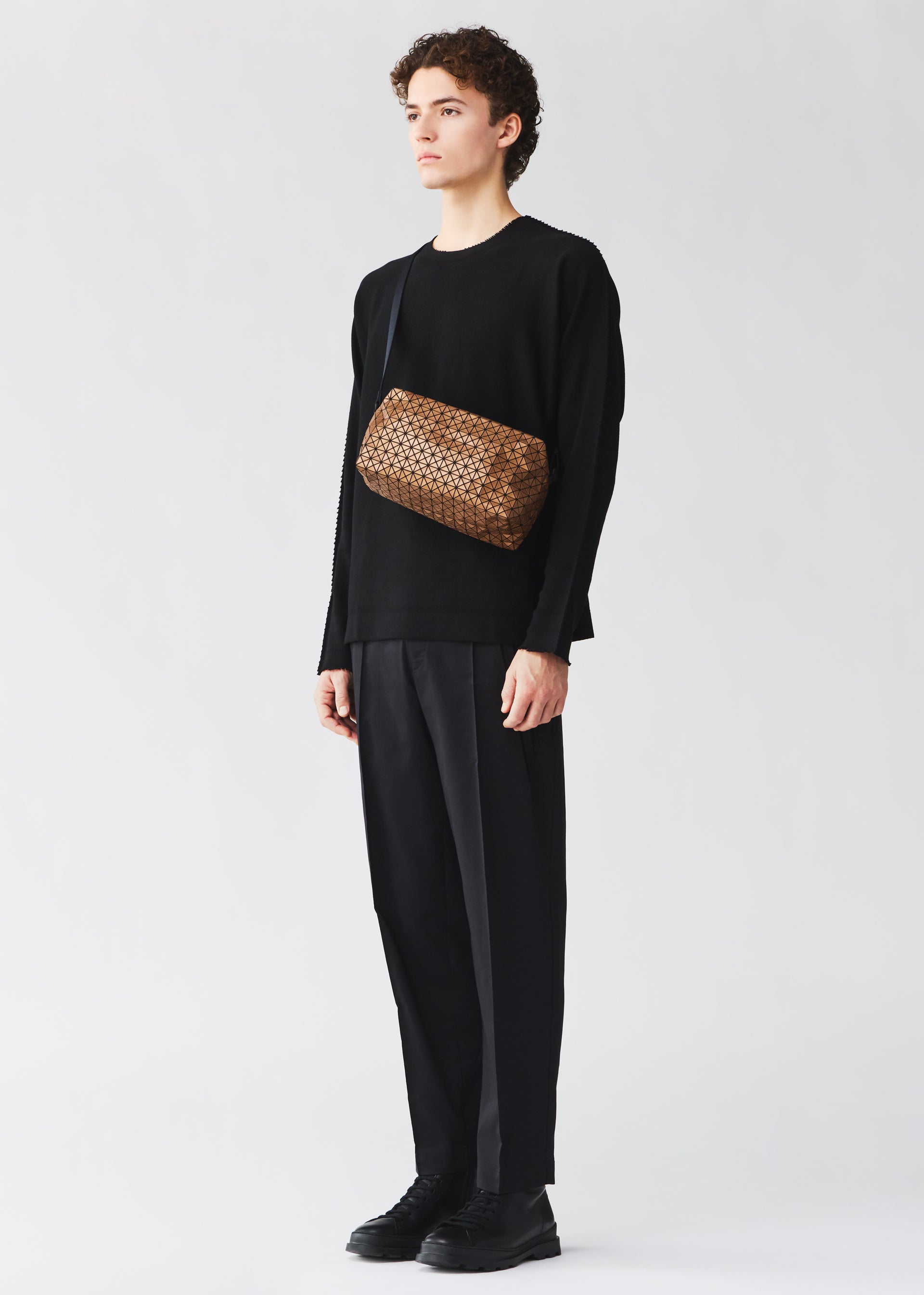 Zoomed view A young person with short curly hair, dressed in all black, stands facing left wearing the BAO BAO ISSEY MIYAKE SADDLE BAG REFLECTOR over one shoulder. The background is plain light gray.