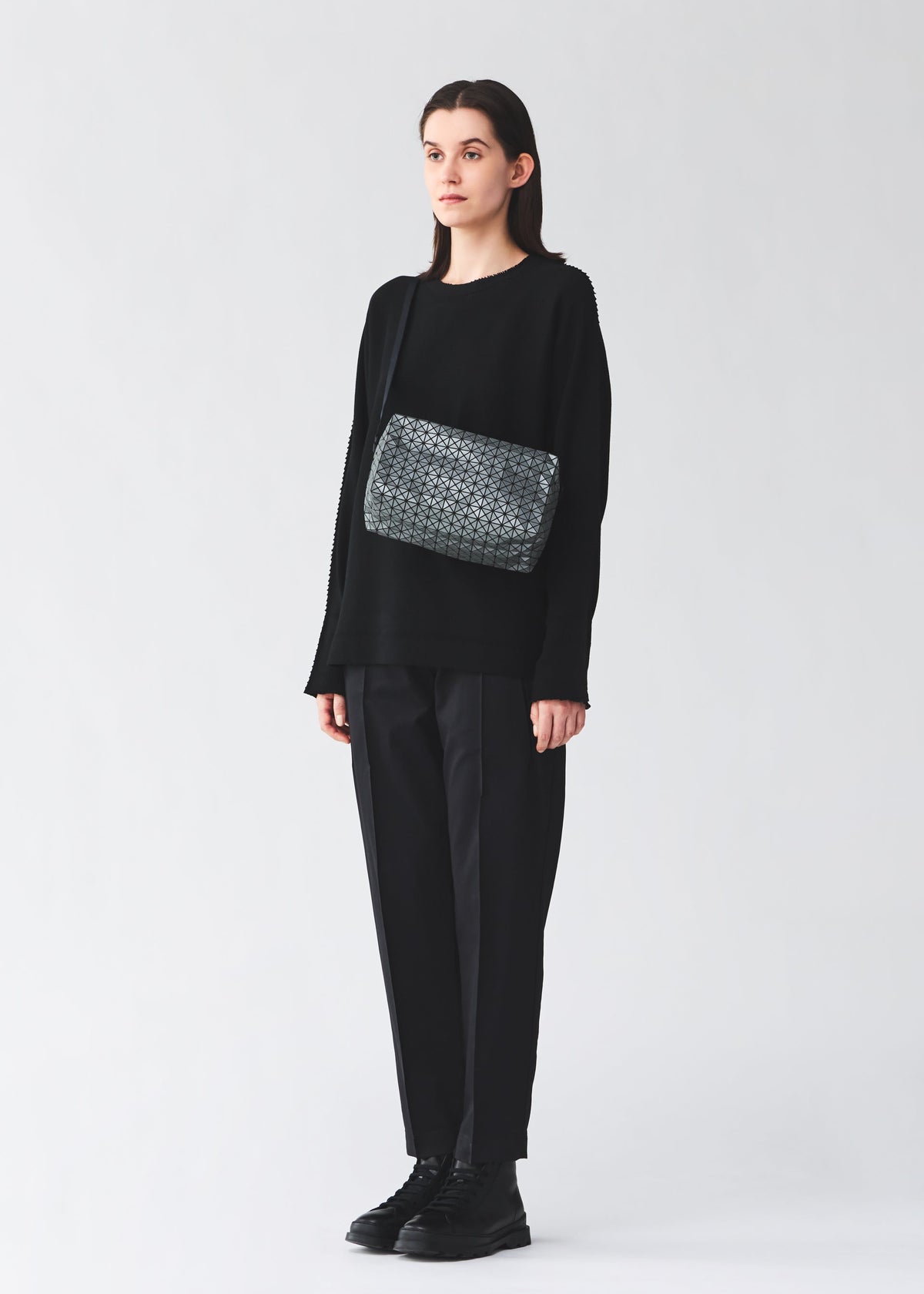 A person with straight dark hair stands against a plain light background, dressed in black and carrying the SADDLE BAG REFLECTOR by BAO BAO ISSEY MIYAKE—a large, reflective shoulder bag. view 7 of 8