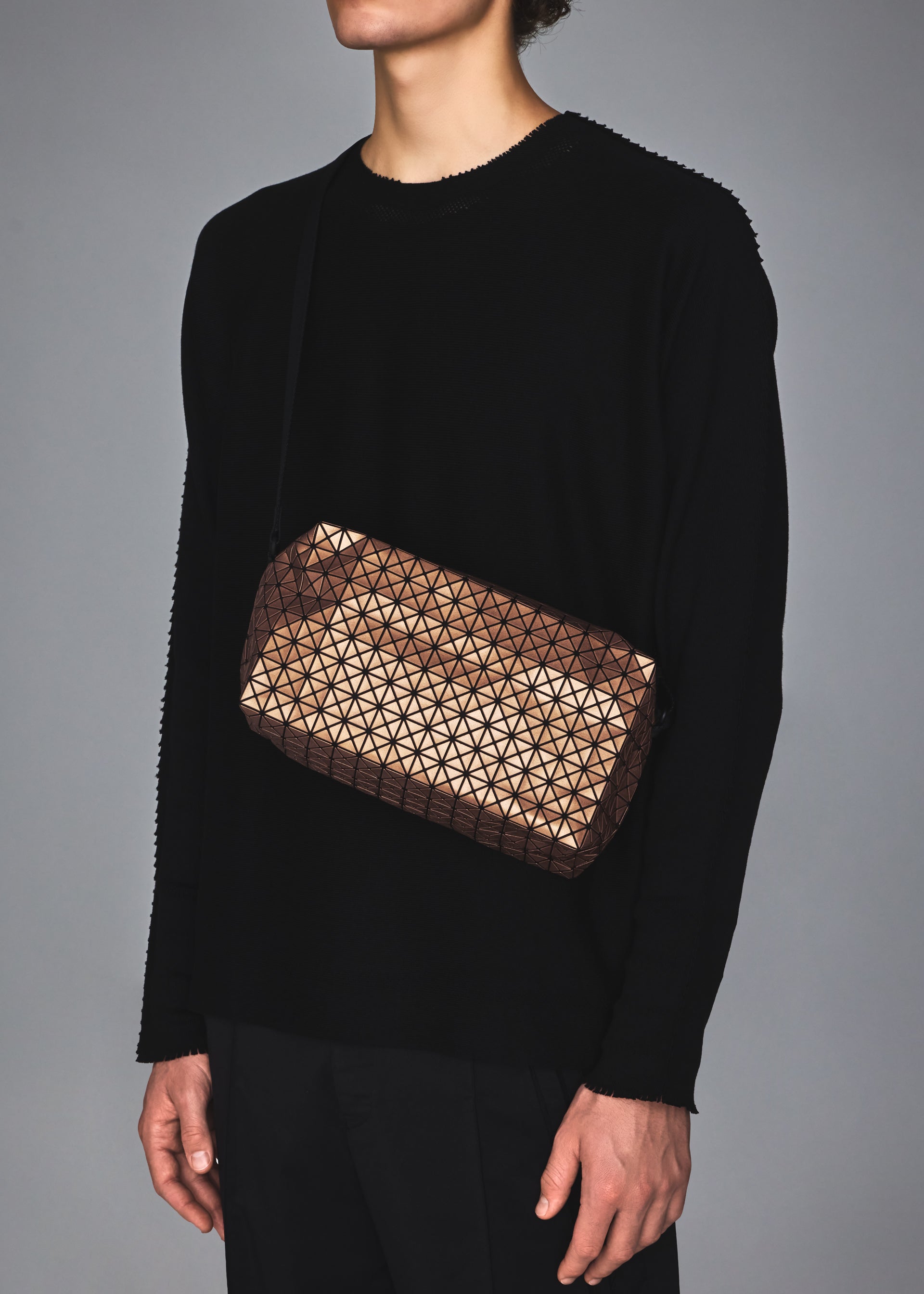 Zoomed view A person in all black is shown from the shoulders down, carrying the BAO BAO ISSEY MIYAKE SADDLE BAG REFLECTOR with a signature triangular pattern, its zippered pocket catching the light against a plain gray background.