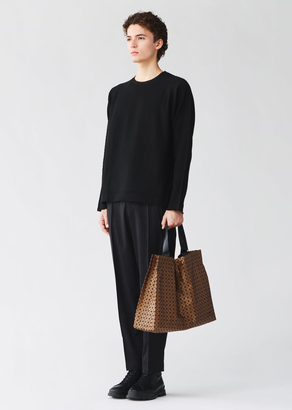 A person with curly hair stands against a plain light background, dressed in all black and holding the BAO BAO ISSEY MIYAKE CART REFLECTOR metallic tote bag with adjustable handles. view 6 of 8