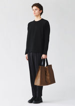 A person with curly hair stands against a plain light background, dressed in all black and holding the BAO BAO ISSEY MIYAKE CART REFLECTOR metallic tote bag with adjustable handles.