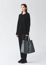 A person in loose black clothes and shoes stands against a plain light background, holding the BAO BAO ISSEY MIYAKE CART REFLECTOR metallic tote bag with adjustable handles by their side.