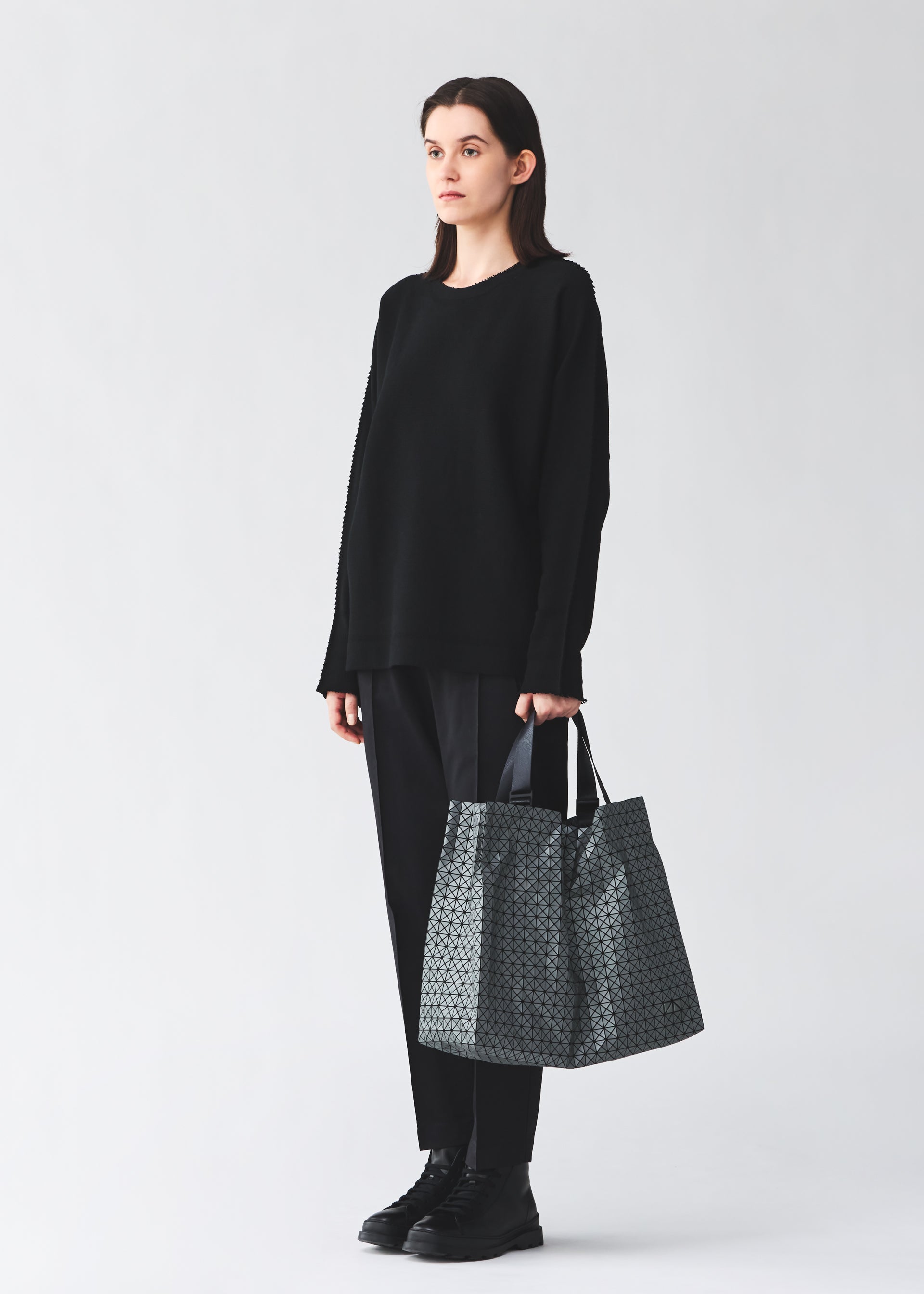 Zoomed view A person in loose black clothes and shoes stands against a plain light background, holding the BAO BAO ISSEY MIYAKE CART REFLECTOR metallic tote bag with adjustable handles by their side.