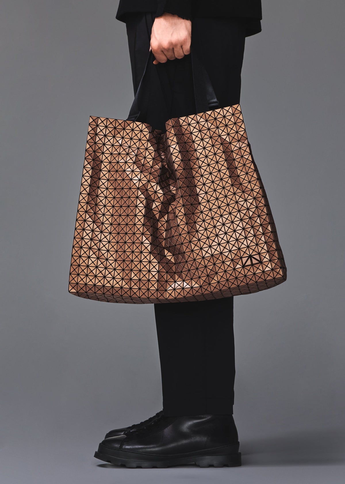 A person dressed in black holds the BAO BAO ISSEY MIYAKE CART REFLECTOR tote, featuring a large geometric pattern and adjustable handles. Only their lower torso, hand, and shoes are visible against a plain gray background. view 8 of 8