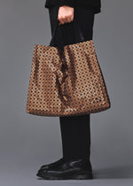 A person dressed in black holds the BAO BAO ISSEY MIYAKE CART REFLECTOR tote, featuring a large geometric pattern and adjustable handles. Only their lower torso, hand, and shoes are visible against a plain gray background.