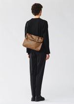 A person with short curly hair in black attire and shoes faces away, carrying the BAO BAO ISSEY MIYAKE FROGGY REFLECTOR crossbody bag with cushioned adjustable straps over their shoulder against a plain light background.