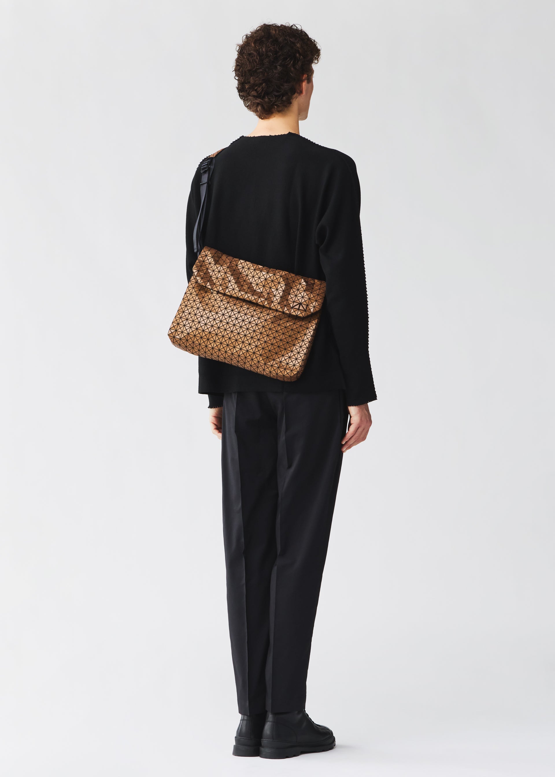 Zoomed view A person with short curly hair in black attire and shoes faces away, carrying the BAO BAO ISSEY MIYAKE FROGGY REFLECTOR crossbody bag with cushioned adjustable straps over their shoulder against a plain light background.