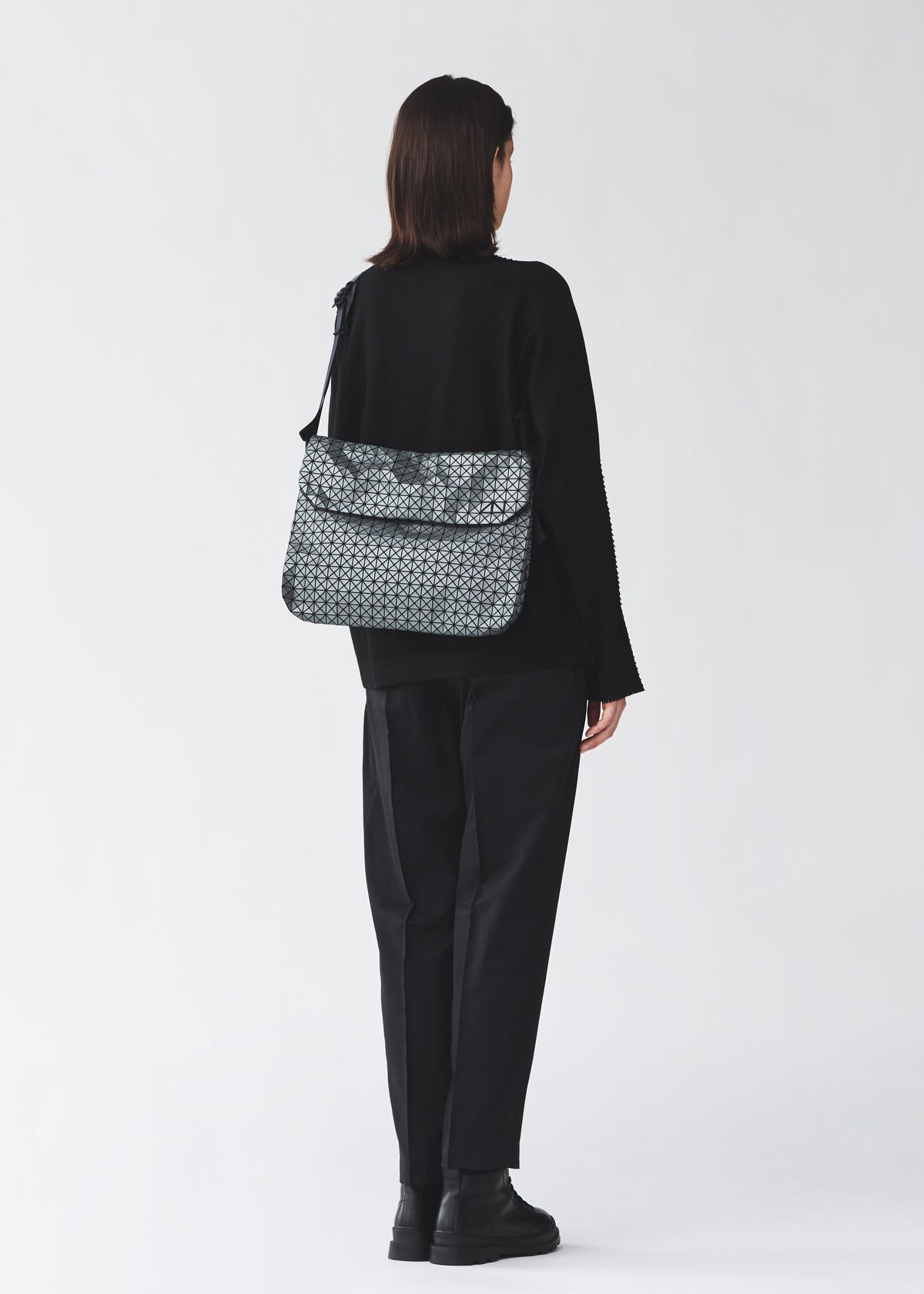 A person with shoulder-length dark hair, in black clothing and boots, stands facing away wearing the BAO BAO ISSEY MIYAKE FROGGY REFLECTOR—a reflective flap bag with cushioned adjustable straps and large storage, against a light background. view 7 of 8