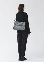 A person with shoulder-length dark hair, in black clothing and boots, stands facing away wearing the BAO BAO ISSEY MIYAKE FROGGY REFLECTOR—a reflective flap bag with cushioned adjustable straps and large storage, against a light background.