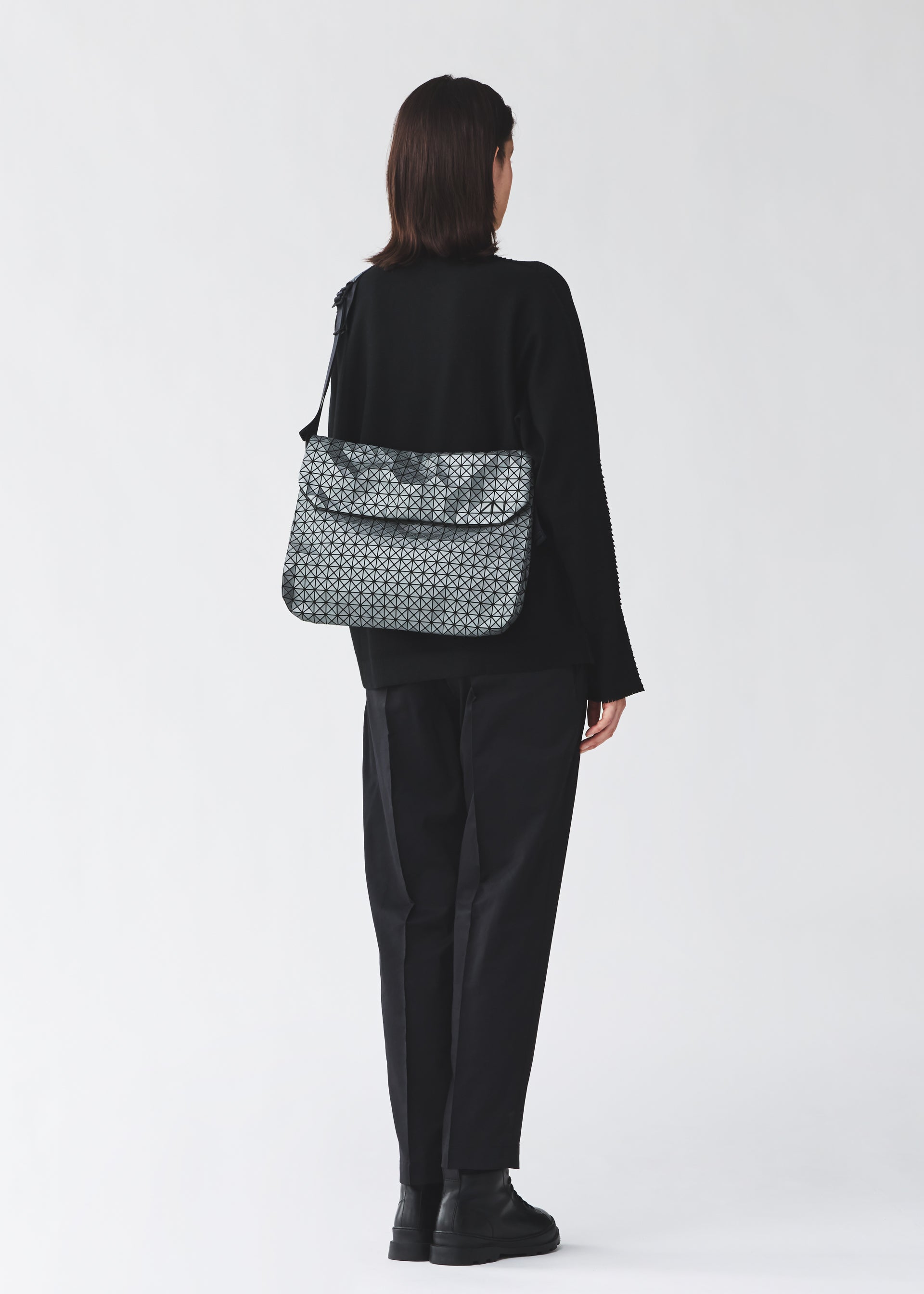 Zoomed view A person with shoulder-length dark hair, in black clothing and boots, stands facing away wearing the BAO BAO ISSEY MIYAKE FROGGY REFLECTOR—a reflective flap bag with cushioned adjustable straps and large storage, against a light background.