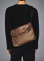 A person wearing a black long-sleeve top and pants carries the BAO BAO ISSEY MIYAKE FROGGY REFLECTOR, a bronze geometric-patterned reflective shoulder bag with a flap, seen from behind.