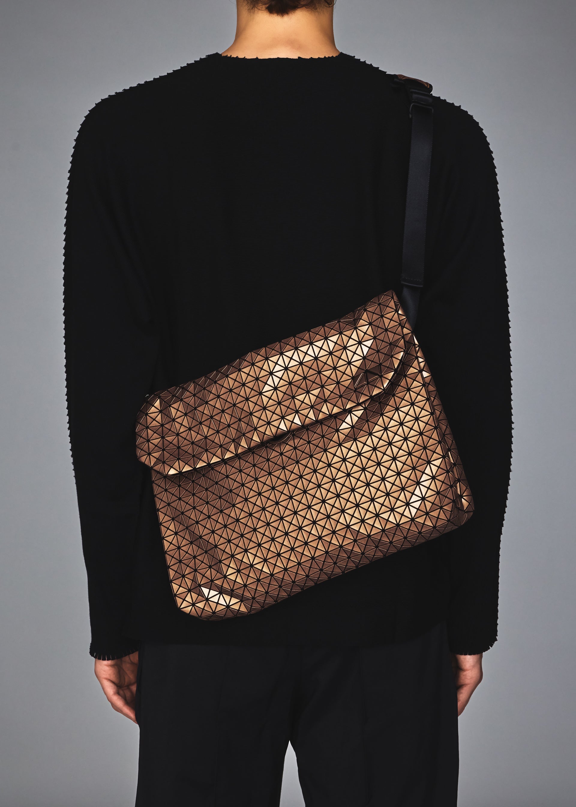 Zoomed view A person wearing a black long-sleeve top and pants carries the BAO BAO ISSEY MIYAKE FROGGY REFLECTOR, a bronze geometric-patterned reflective shoulder bag with a flap, seen from behind.
