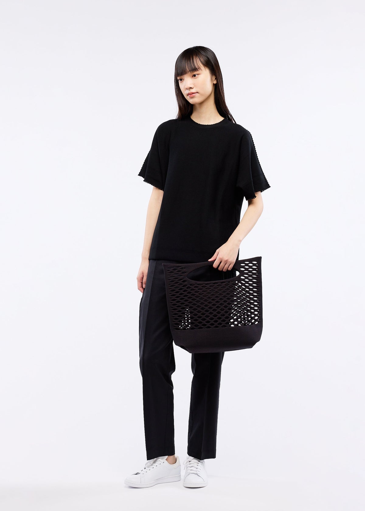 A woman with long dark hair stands against a white background, wearing black pants, a loose black t-shirt, and white sneakers. She holds the GOOD GOODS ISSEY MIYAKE MOKKO BASKET bag in one hand. view 5 of 8
