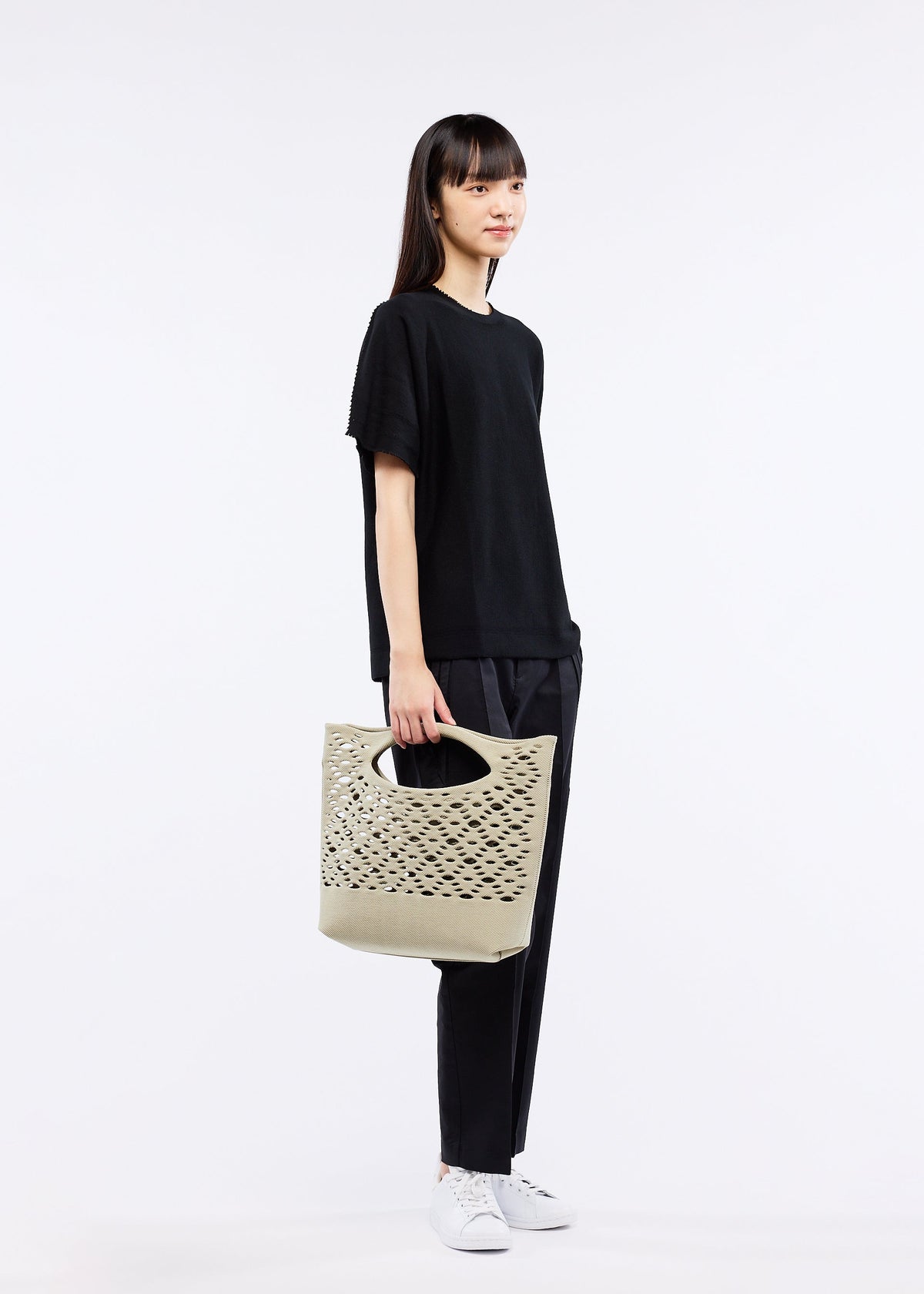A woman with straight dark hair, dressed in a loose black t-shirt, black pants, and white sneakers, stands against a plain white background holding the MOKKO BASKET by GOOD GOODS ISSEY MIYAKE—a light beige tote with laser-cut details. view 6 of 8