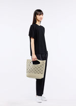 A woman with straight dark hair, dressed in a loose black t-shirt, black pants, and white sneakers, stands against a plain white background holding the MOKKO BASKET by GOOD GOODS ISSEY MIYAKE—a light beige tote with laser-cut details.