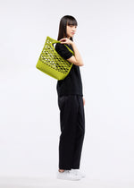 A person with long, straight dark hair stands in profile against a plain white background, dressed in black and white sneakers, carrying the bright green GOOD GOODS ISSEY MIYAKE MOKKO BASKET tote bag with a twill-like lattice pattern over their shoulder.