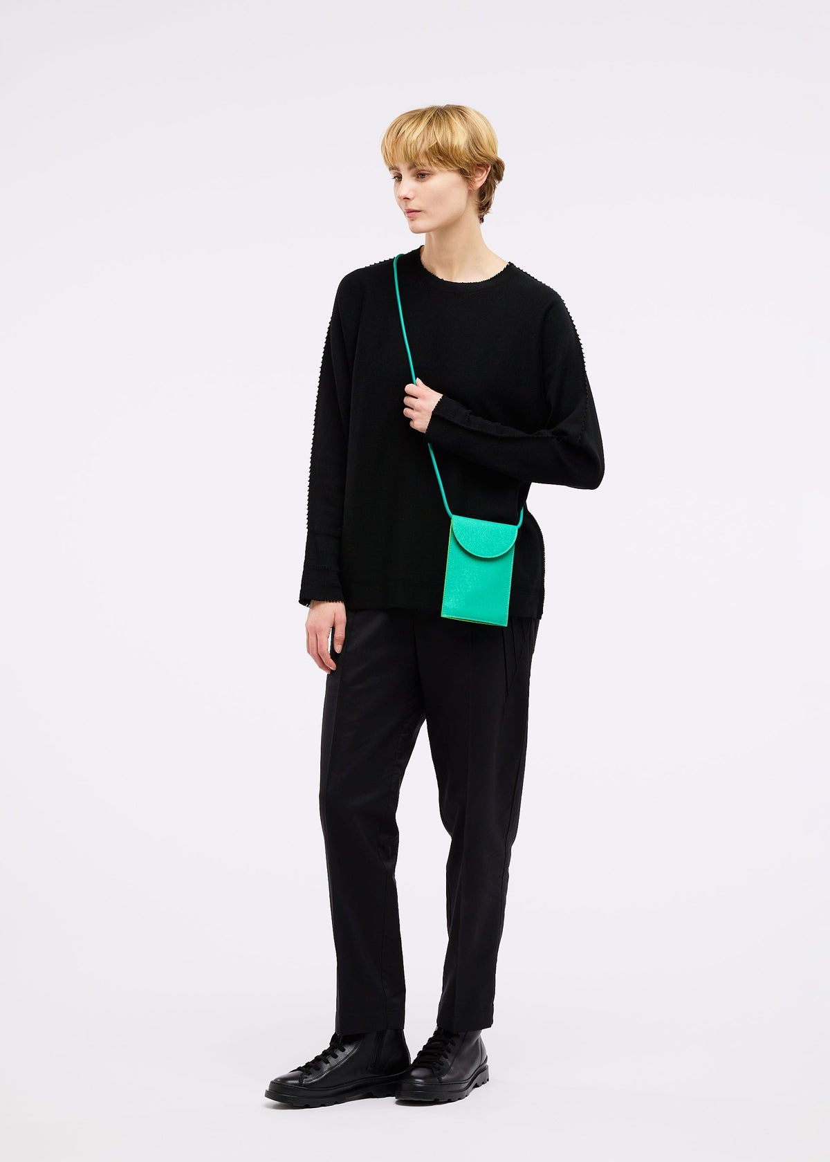 A person with short blond hair stands against a white background wearing black clothing and carries the PACKAGE pochette by GOOD GOODS ISSEY MIYAKE in bright green, resembling a charming gift. view 8 of 9