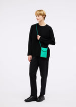 A person with short blond hair stands against a white background wearing black clothing and carries the PACKAGE pochette by GOOD GOODS ISSEY MIYAKE in bright green, resembling a charming gift.