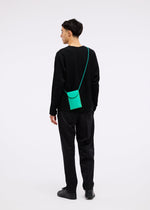 A man in black pants and a black long-sleeve shirt faces away, wearing the small, bright green PACKAGE pochette by GOOD GOODS ISSEY MIYAKE against a plain white background.
