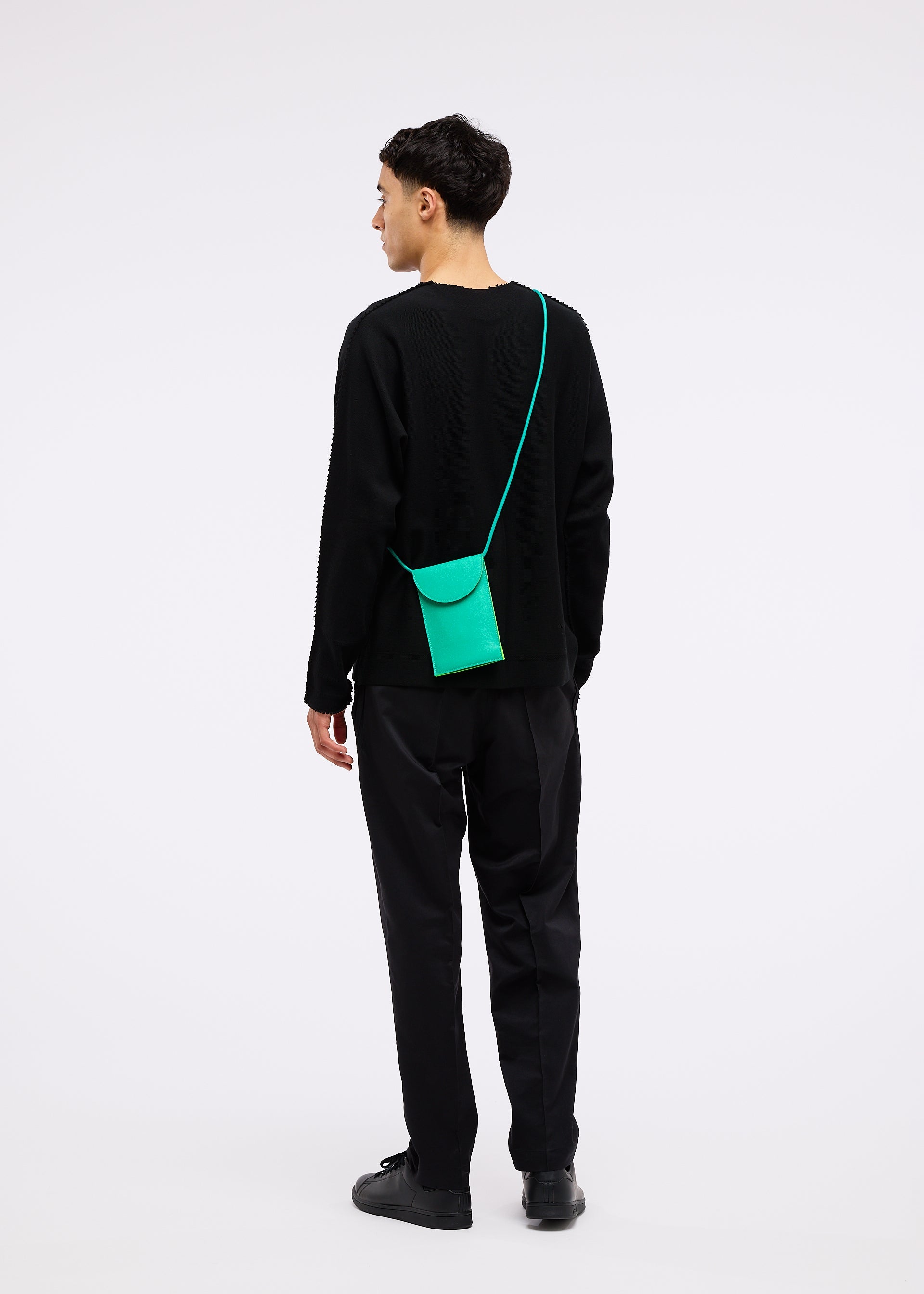 Zoomed view A man in black pants and a black long-sleeve shirt faces away, wearing the small, bright green PACKAGE pochette by GOOD GOODS ISSEY MIYAKE against a plain white background.