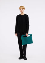 A person with short blonde hair stands against a white background wearing black clothes and holding the KURURI large teal fabric bag with an adjustable handle and cutout detail by GOOD GOODS ISSEY MIYAKE.