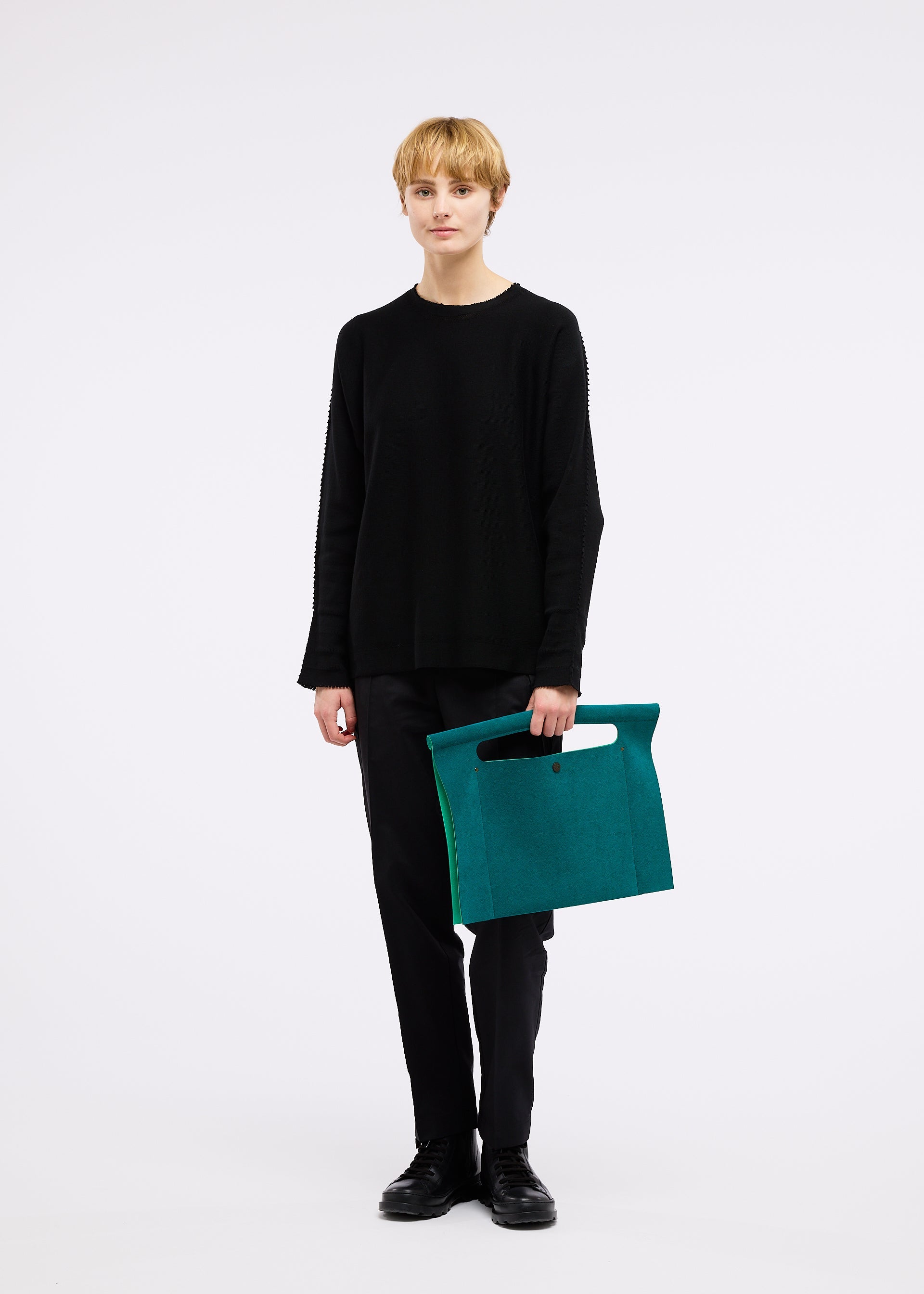 Zoomed view A person with short blonde hair stands against a white background wearing black clothes and holding the KURURI large teal fabric bag with an adjustable handle and cutout detail by GOOD GOODS ISSEY MIYAKE.