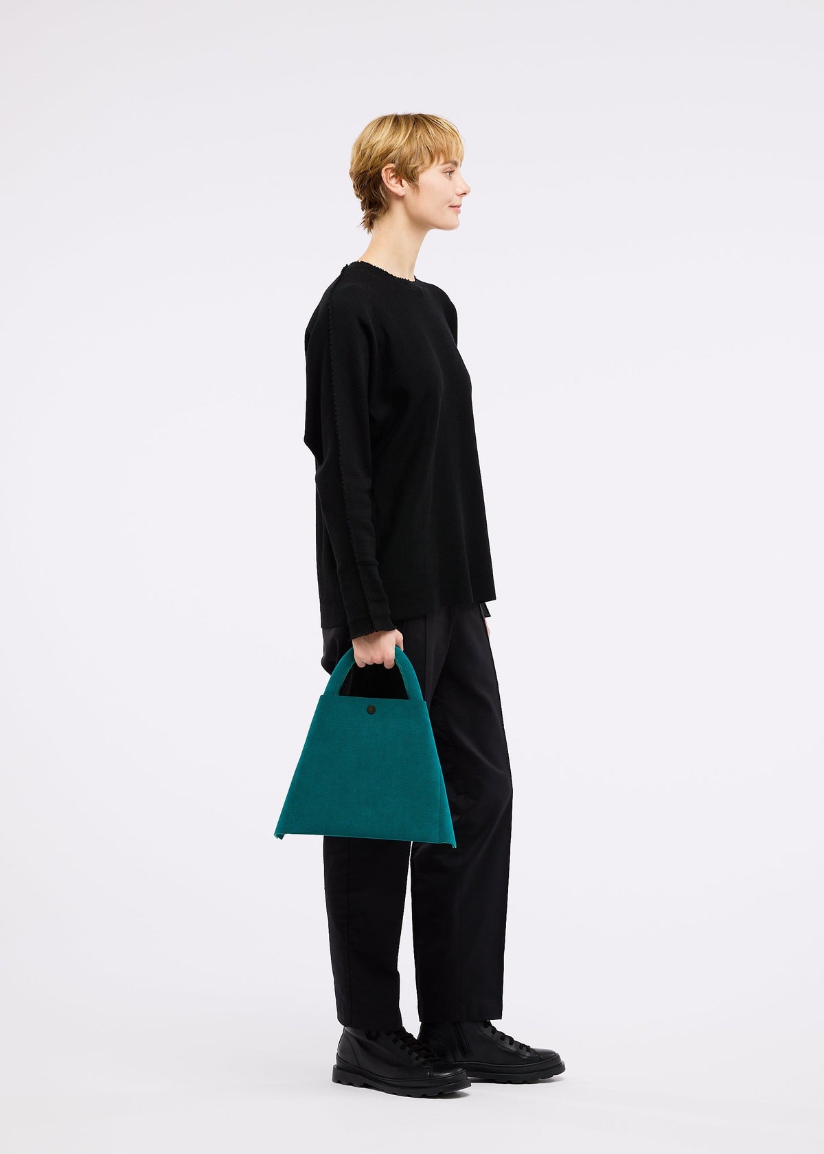 A person in a loose black sweater and pants stands in profile holding the KURURI teal fabric bag with adjustable handle by GOOD GOODS ISSEY MIYAKE, set against a plain white background. view 15 of 15