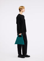 A person in a loose black sweater and pants stands in profile holding the KURURI teal fabric bag with adjustable handle by GOOD GOODS ISSEY MIYAKE, set against a plain white background.