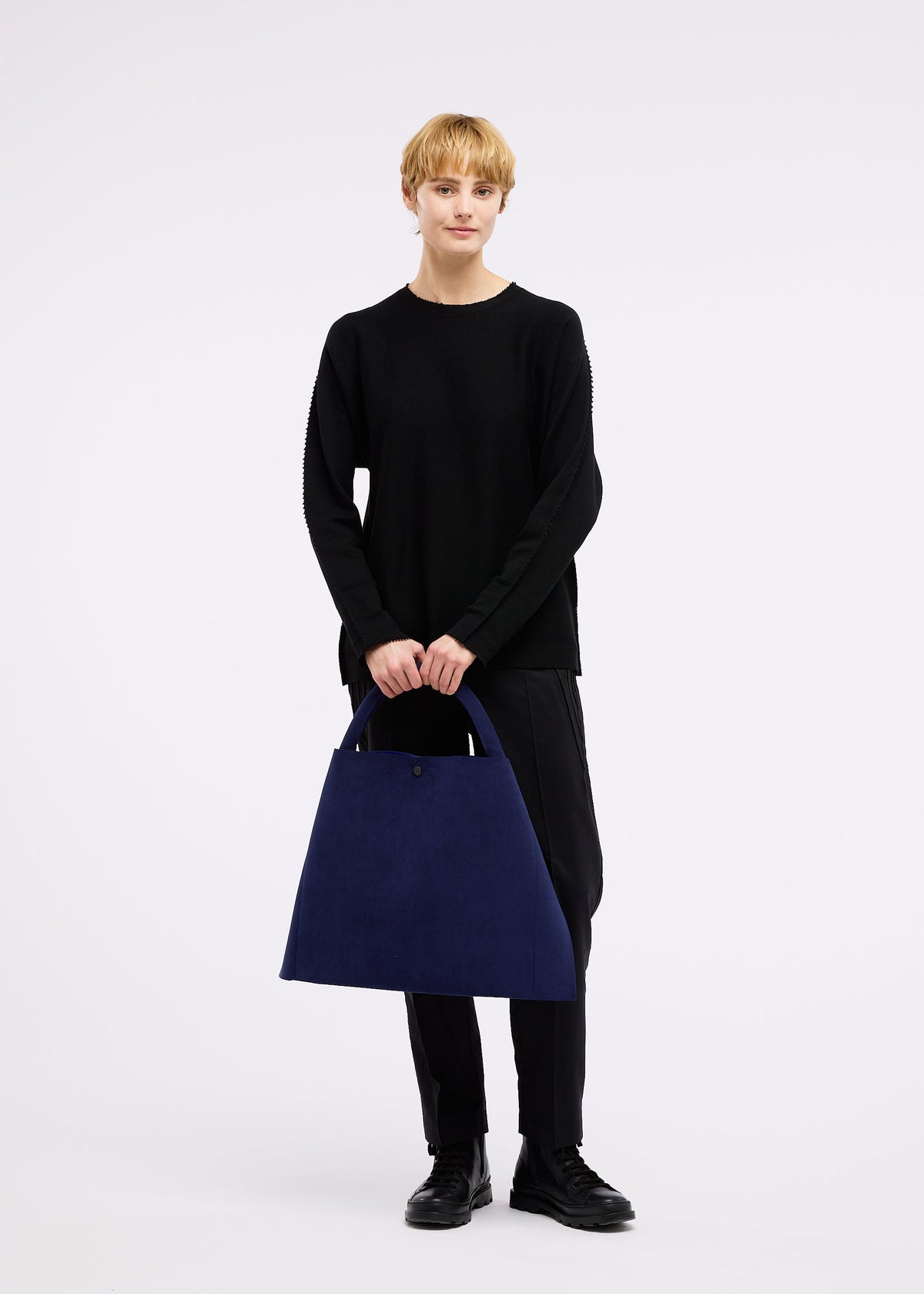 A person with short blonde hair in black long sleeves and pants holds the large blue KURURI winding bag by GOOD GOODS ISSEY MIYAKE, featuring a strong handle. The background is plain white. view 14 of 17