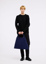A person with short blonde hair in black long sleeves and pants holds the large blue KURURI winding bag by GOOD GOODS ISSEY MIYAKE, featuring a strong handle. The background is plain white.