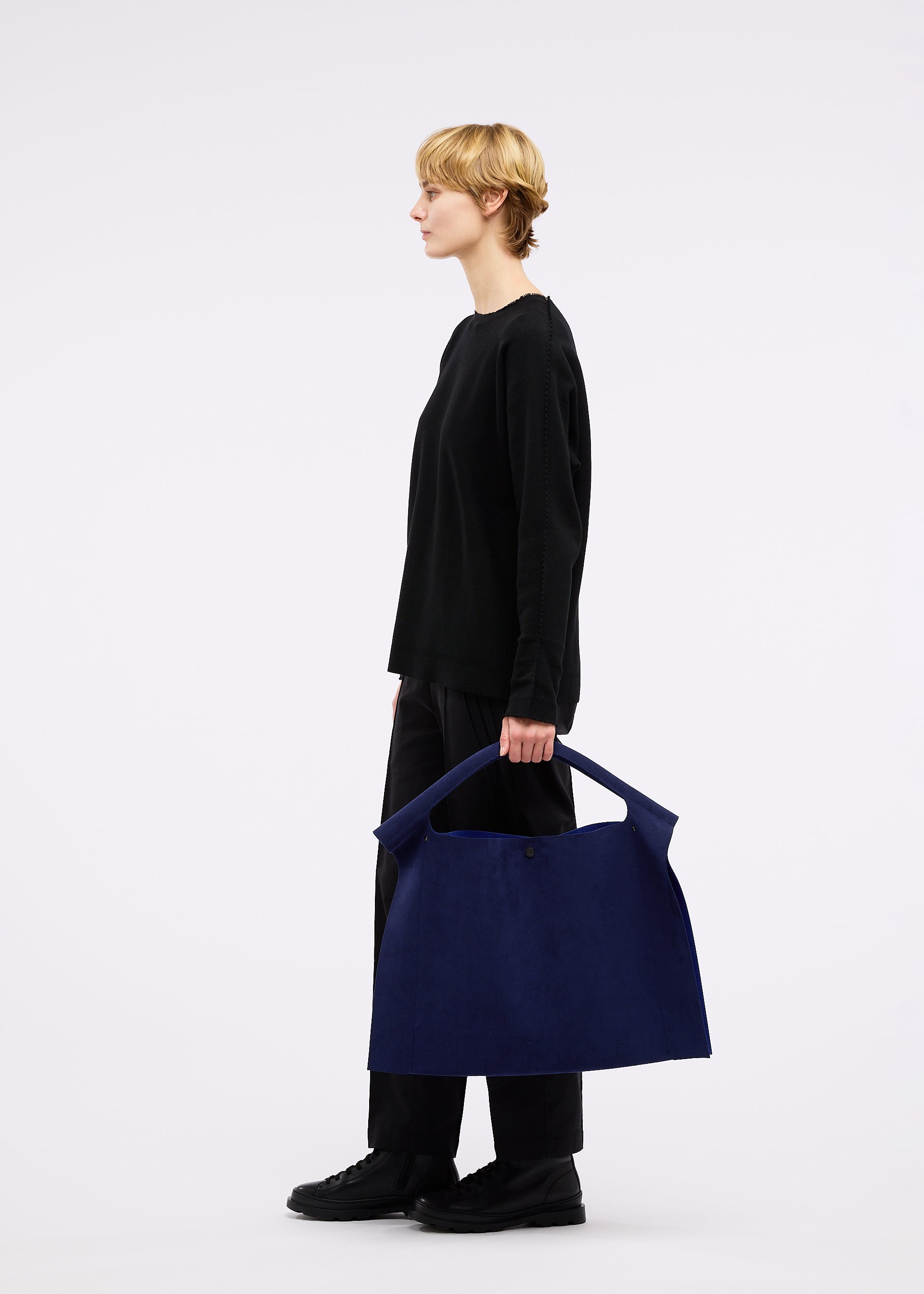 Zoomed view A person with short blond hair, dressed in black, stands in profile against a white background holding the KURURI bag by GOOD GOODS ISSEY MIYAKE—a large dark blue adjustable silhouette bag with a strong handle.