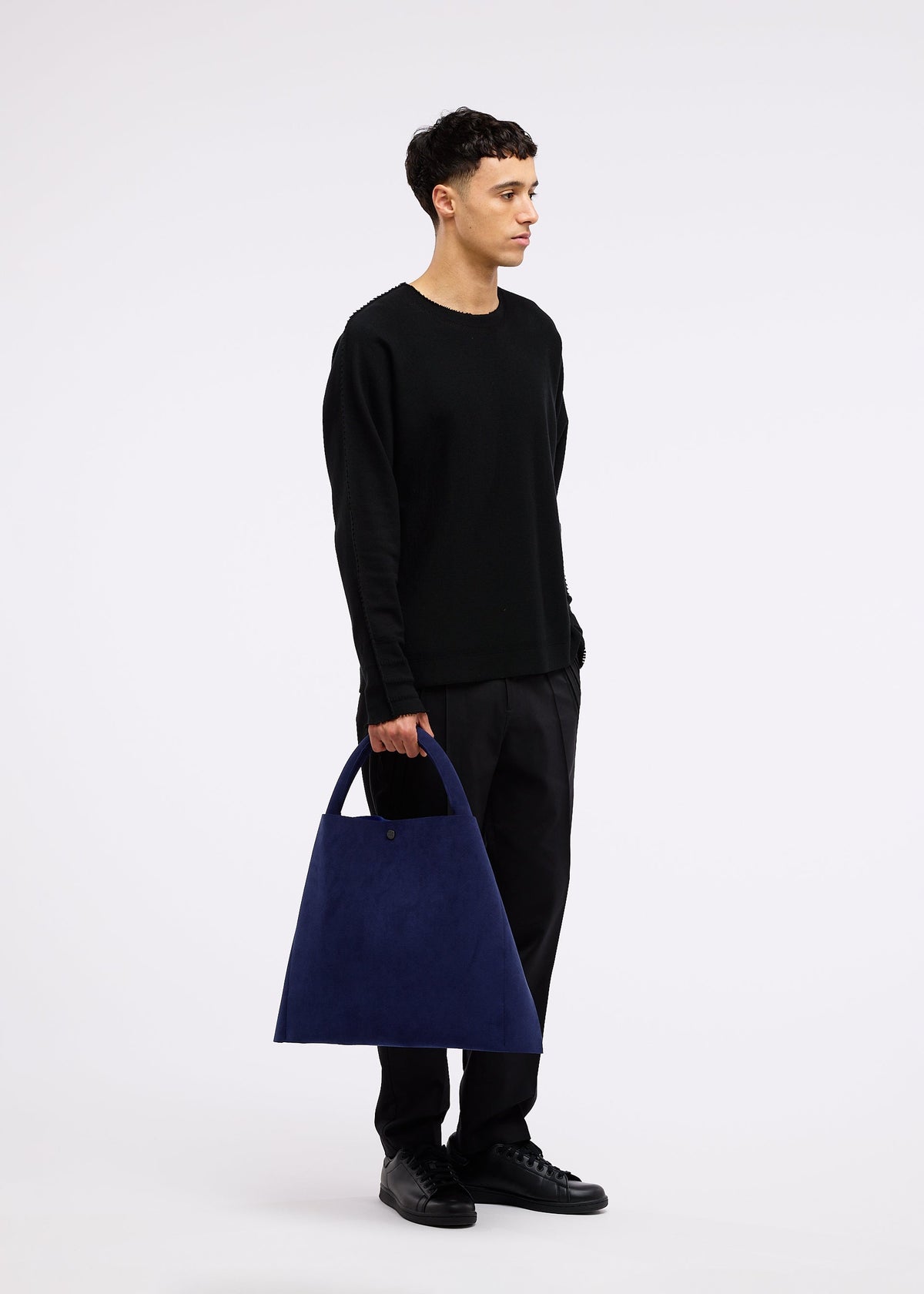 A person in black stands against a white background, holding the large, triangular navy KURURI adjustable silhouette bag by GOOD GOODS ISSEY MIYAKE with a strong handle at their side. view 16 of 17