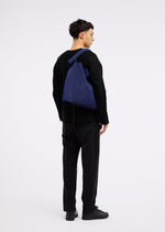A man in all-black attire and shoes faces left, carrying the KURURI blue adjustable silhouette bag by GOOD GOODS ISSEY MIYAKE across his back. The background is plain white.