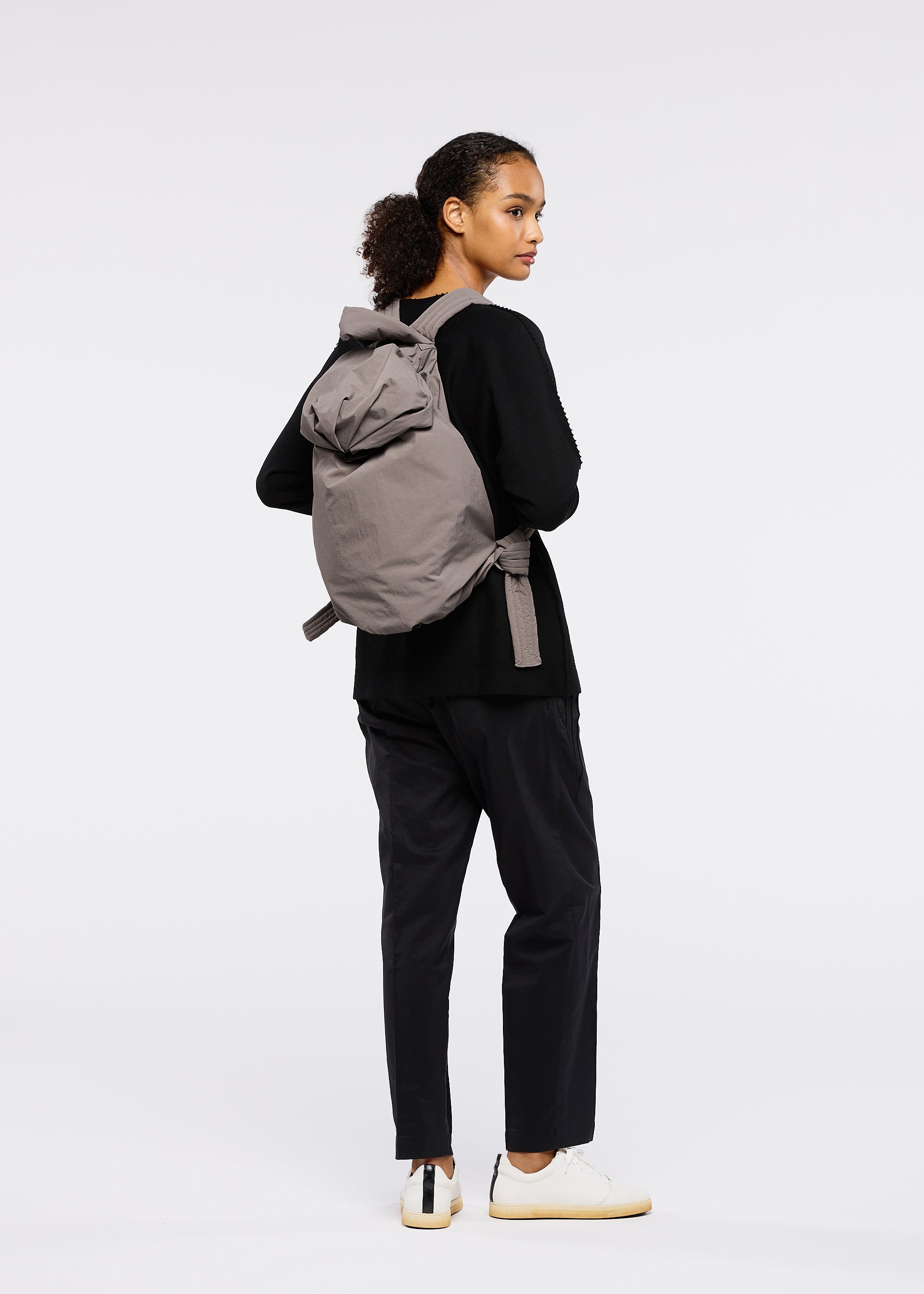 Zoomed view A person stands against a plain white background, looking over their shoulder, wearing the FUWA FUWA gray zero-waste backpack by GOOD GOODS ISSEY MIYAKE with a black long-sleeve top, black pants, and white sneakers.