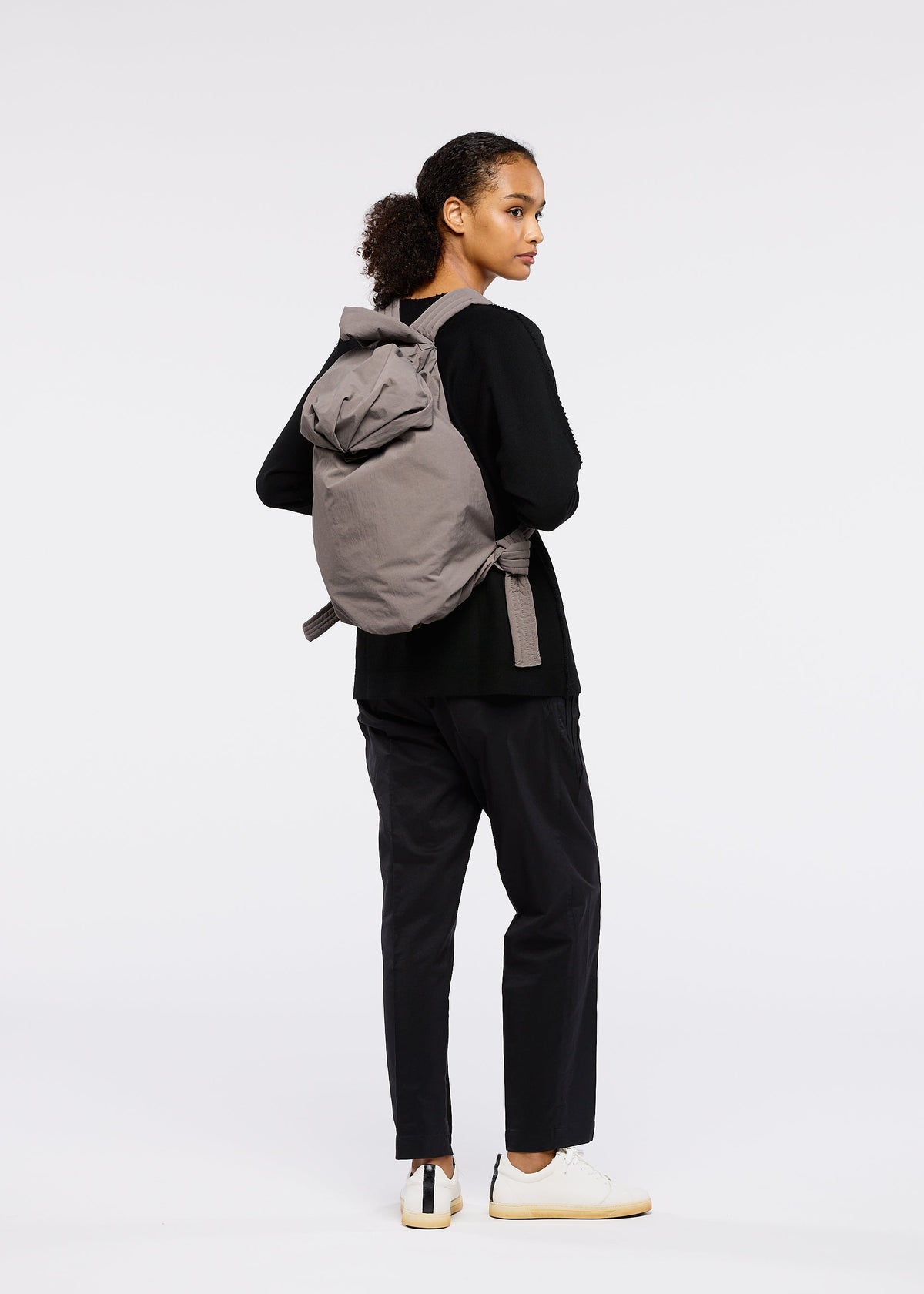 A woman with curly hair in a ponytail stands slightly turned, dressed in black noragi-inspired attire, white sneakers, and carries the gray FUWA FUWA backpack by GOOD GOODS ISSEY MIYAKE against a plain white background. view 6 of 7