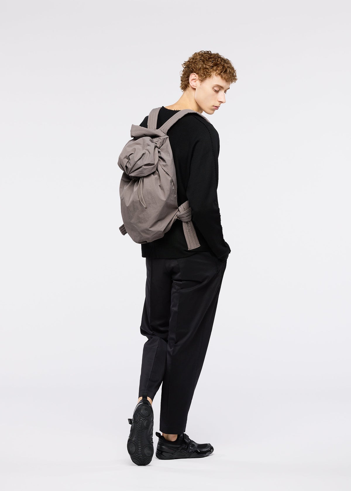 A person with curly hair models the FUWA FUWA backpack by GOOD GOODS ISSEY MIYAKE, wearing black Japanese workwear-inspired pants and a long-sleeve shirt, standing sideways against a plain white background. view 7 of 7