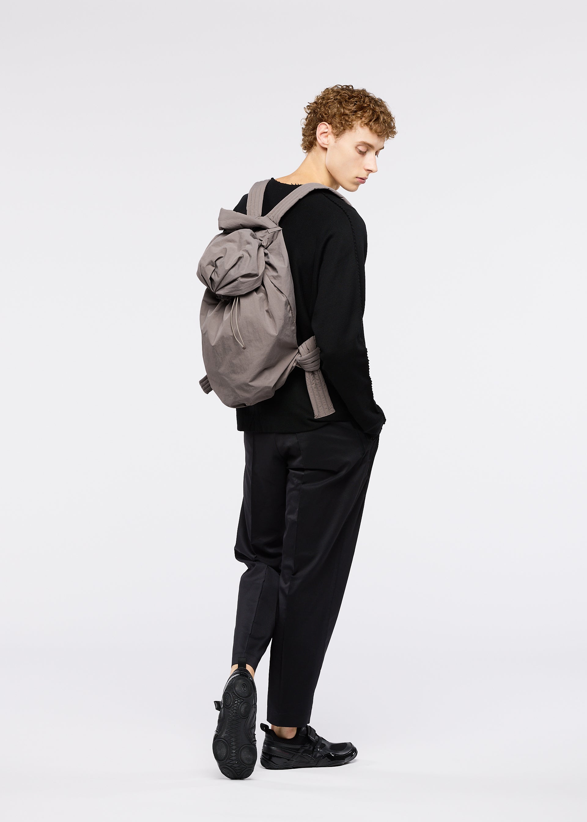 Zoomed view A person with curly hair stands sideways against a white background, wearing all black and carrying the FUWA FUWA backpack by GOOD GOODS ISSEY MIYAKE, looking down.