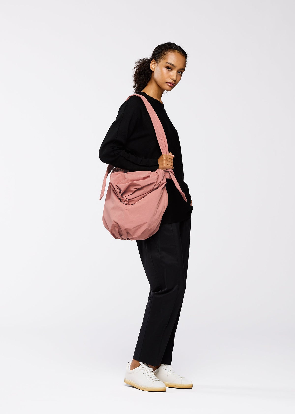A woman stands against a white background, wearing black clothes and white sneakers. She carries the FUWA FUWA large pink zero-waste shoulder bag by GOOD GOODS ISSEY MIYAKE, looking slightly to the side with a neutral expression. view 6 of 7