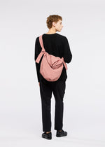 A person with short curly hair, in a black long-sleeve top and pants, stands facing away against a white background carrying the FUWA FUWA large light pink shoulder bag by GOOD GOODS ISSEY MIYAKE, inspired by zero-waste design.