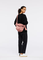 A person with curly hair stands sideways, looking back at the camera. They wear FUWA FUWA black pants by GOOD GOODS ISSEY MIYAKE, a black top, white sneakers, and a pink shoulder bag against a plain light background.