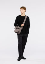 A person with short curly hair stands against a white background, wearing black clothes and shoes, carrying the FUWA FUWA shoulder bag by GOOD GOODS ISSEY MIYAKE.