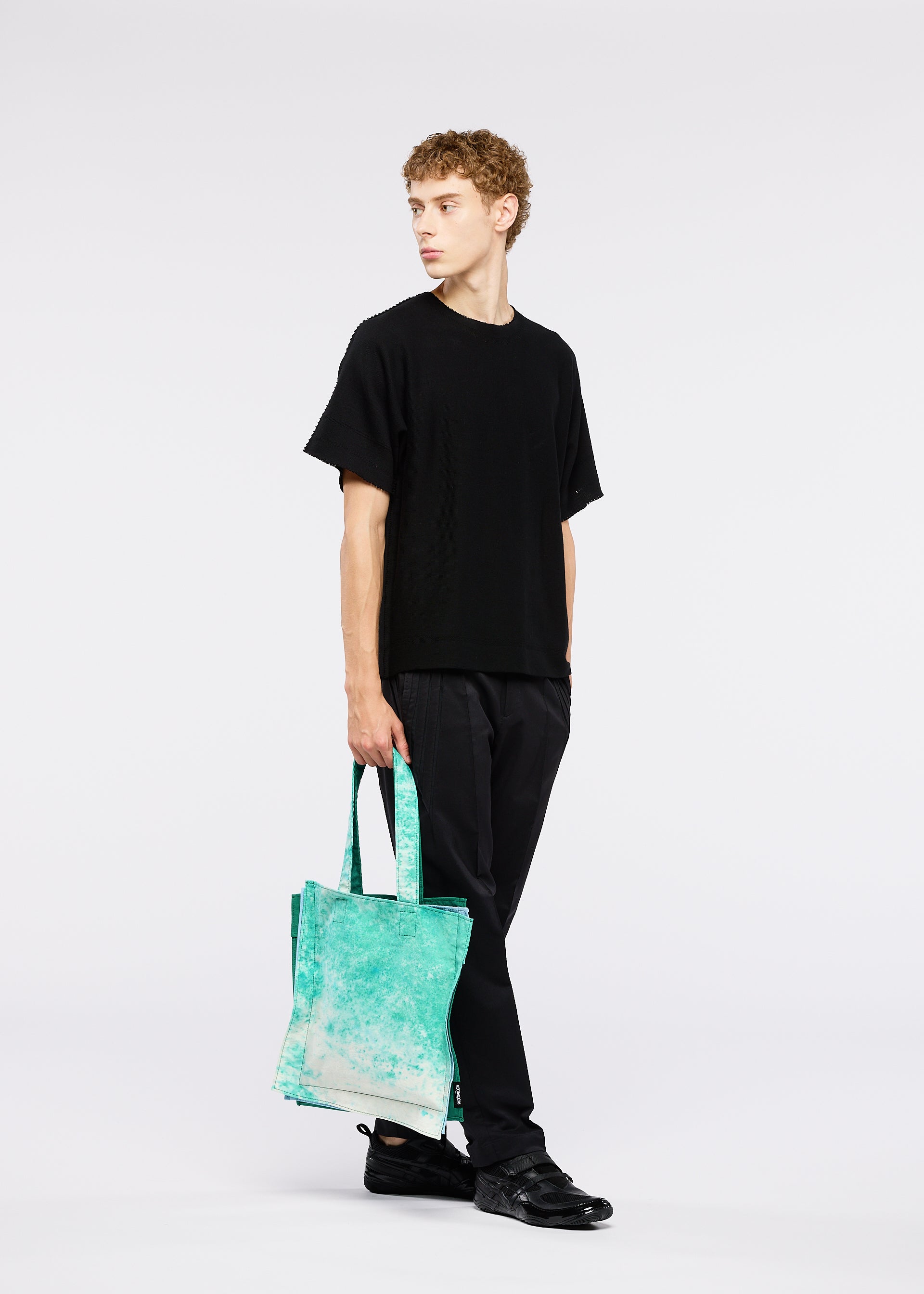 Zoomed view A young man with curly hair wears all black and holds a GOOD GOODS ISSEY MIYAKE IROGAMI SALT DYE tote bag in turquoise, standing against a plain white background.