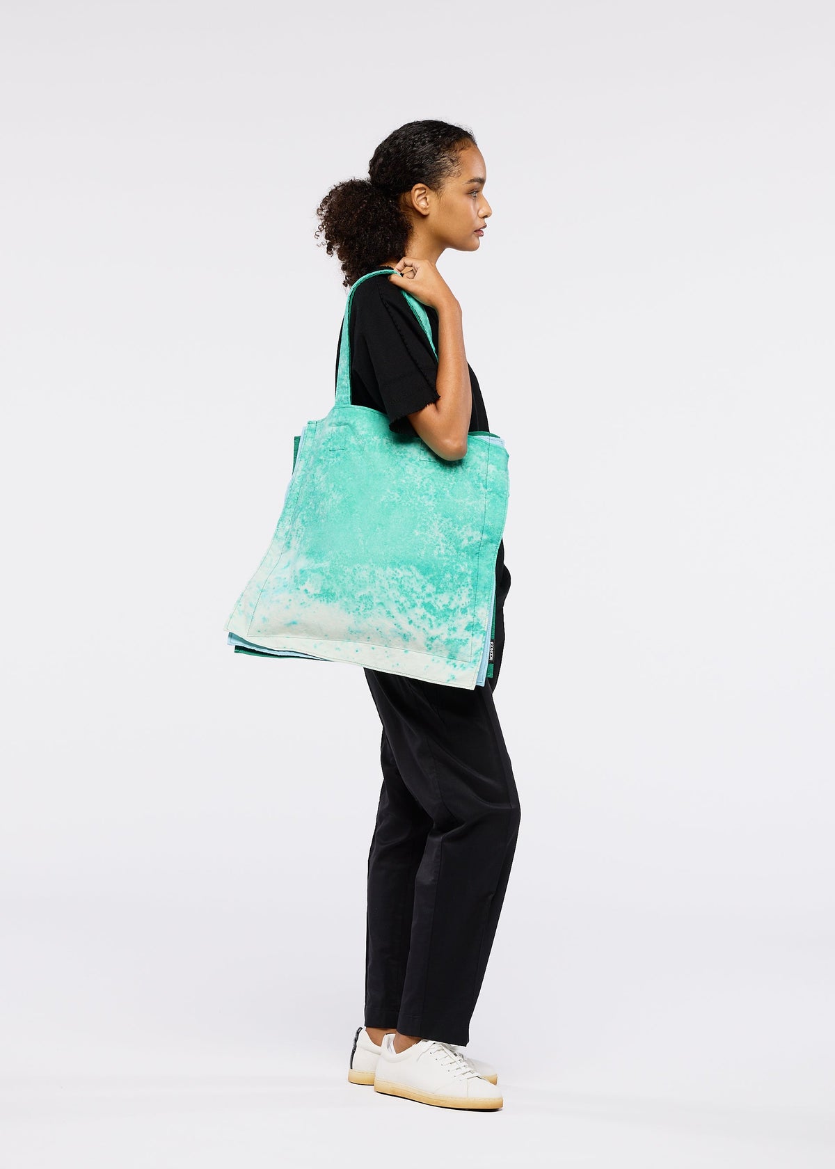 A woman in black pants, a black t-shirt, and white sneakers stands in profile against a white background, carrying the large turquoise IROGAMI SALT DYE tote bag by GOOD GOODS ISSEY MIYAKE over her shoulder. view 9 of 10
